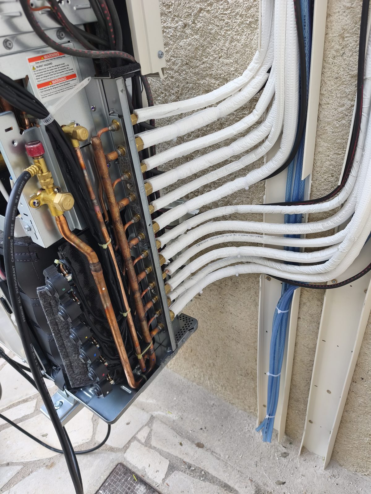 Groupe extérieur ouvert, douze tuyaux isolés blancs alignés sur leurs raccords en laiton, tuyaux bleus des condensats dans la goulotte ouverte — MSC Climatisation